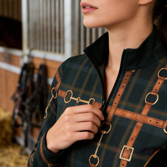 Warm indoor barn close‑up shot focusing on zipper and print detail of female equestrian wearing dark green tartan long‑sleeve quarter‑zip riding top with leather‑strap bridle hardware print