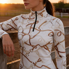 Sunset pasture fence lifestyle shot of female equestrian wearing cream bridle bit and reins print long‑sleeve riding top