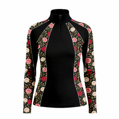 Lifestyle shot outside white stable farmhouse of female rider wearing black vintage rose floral long‑sleeve riding top with rhinestone‑dot panel trim, paired with beige riding breeches