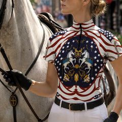 Outdoor portrait of female rider in black gloves holding horse reins wearing patriotic flag‑themed heraldic horse print short‑sleeve riding top beside grey horse