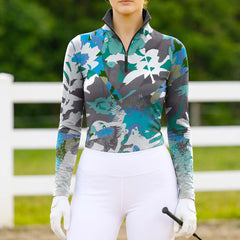 Model wearing the ocean palette version of the Abstract Watercolor Floral Print Long-Sleeve Riding Top at an outdoor equestrian arena, with jumps in the background
