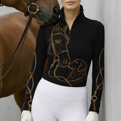Model standing beside brown horse’s head wearing black gold line art horse & golden retriever long sleeve riding top against light grey stable wall
