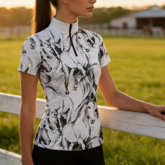 Golden‑hour pasture lifestyle side shot of female equestrian leaning on wooden fence wearing white short‑sleeve quarter‑zip riding top with all‑over black sketch galloping horse print