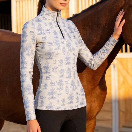 Warm sunlit stable shot of model gently touching a brown horse while wearing cream soft blue toile de jouy horse print long sleeve riding top
