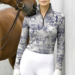 Stable lifestyle shot of female rider beside horse wearing blue toile de jouy woodland‑scene‑print long‑sleeve quarter‑zip riding top