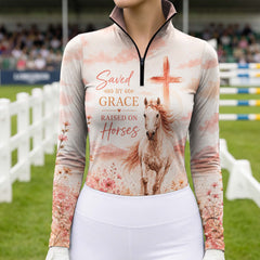 Model wearing warm blush watercolor meadow flower cross horse long sleeve riding top at outdoor equestrian show jumping arena with spectator stands
