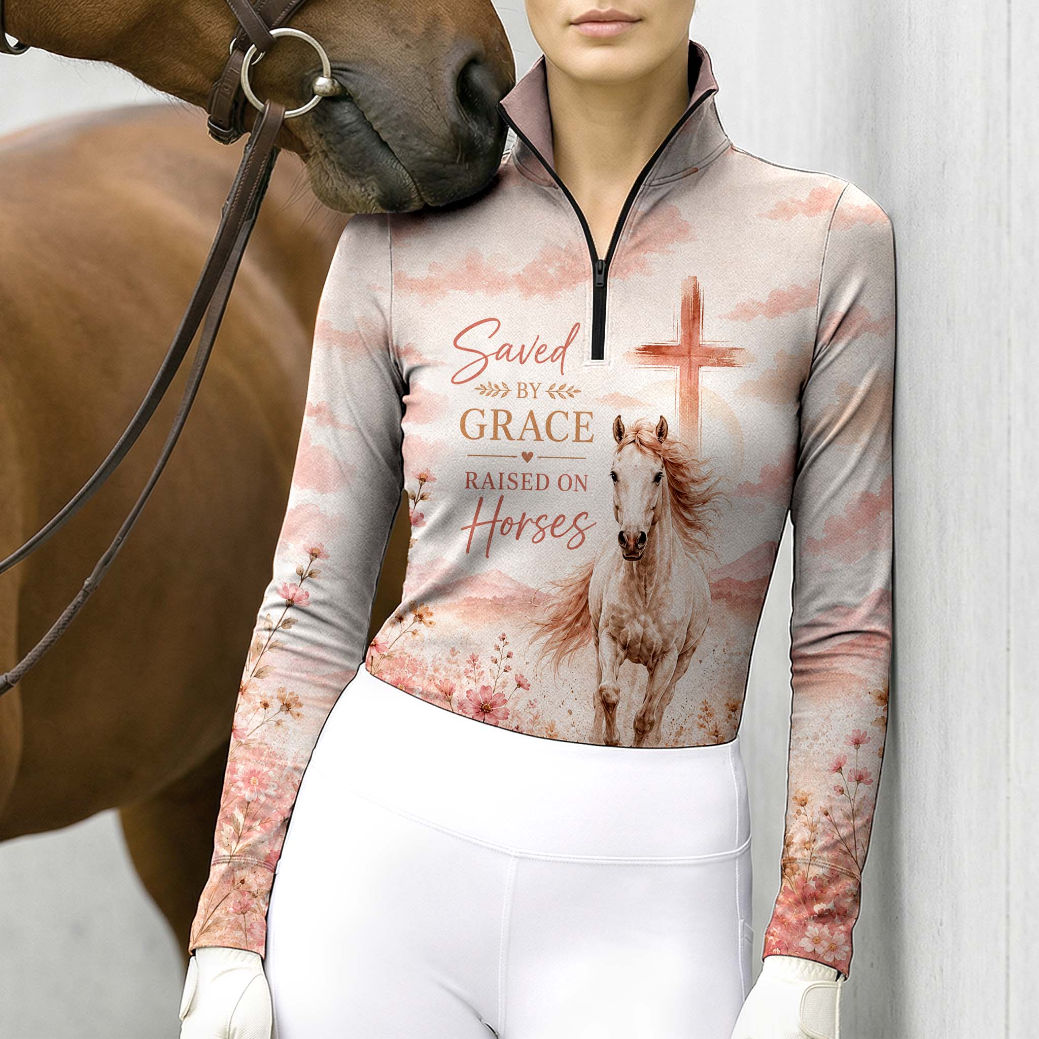 Model standing beside brown horse’s head wearing muted blush galloping horse faith wildflower long sleeve riding top against light grey stable wall

