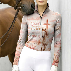 Model standing beside brown horse’s head wearing muted blush galloping horse faith wildflower long sleeve riding top against light grey stable wall
