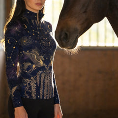 Warm indoor barn shot of female rider beside horse wearing midnight navy celestial pegasus galaxy graphic long‑sleeve riding top