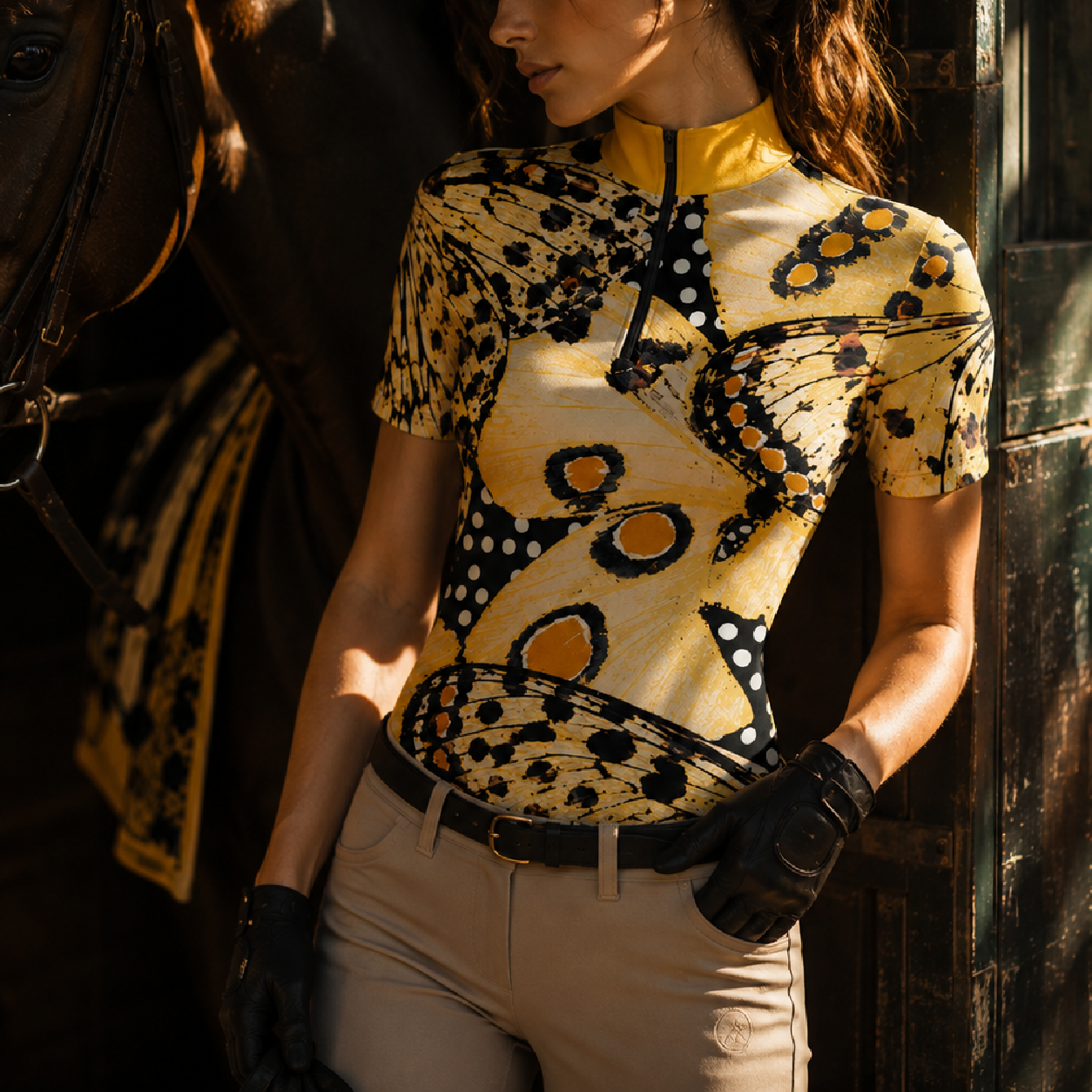 Dappled sunlit stable lifestyle side portrait of female equestrian in black leather gloves beside dark horse wearing golden‑collar vintage moth butterfly polka‑dot print short‑sleeve riding top
