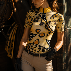 Dappled sunlit stable lifestyle side portrait of female equestrian in black leather gloves beside dark horse wearing golden‑collar vintage moth butterfly polka‑dot print short‑sleeve riding top