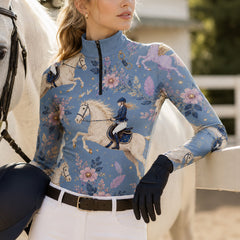 White fence paddock lifestyle shot of female equestrian leaning on fence beside white horse wearing dusty‑blue fairytale pegasus print long‑sleeve riding top, paired with white breeches and black gloves