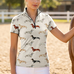 Sand arena lifestyle photo of model wearing cream watercolor dachshund all-over print short sleeve quarter-zip riding top beside horse