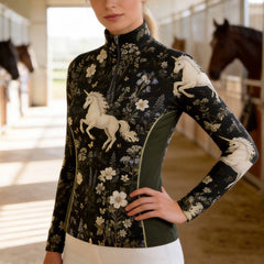 Sunlit equestrian stable walkway shot of model posing wearing dark wildflower white unicorn printed long sleeve quarter-zip riding top with olive contrast panels