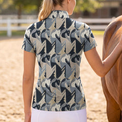 Bright sunlit stable yard shot of female rider viewed from the back standing beside brown horse wearing charcoal sand abstract geometric horse head all-over print short sleeve quarter-zip riding top paired with white riding leggings