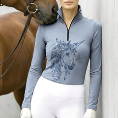 Model gently standing beside brown horse’s head wearing dusty blue sketch feather unicorn long sleeve riding top against light grey stable wall
