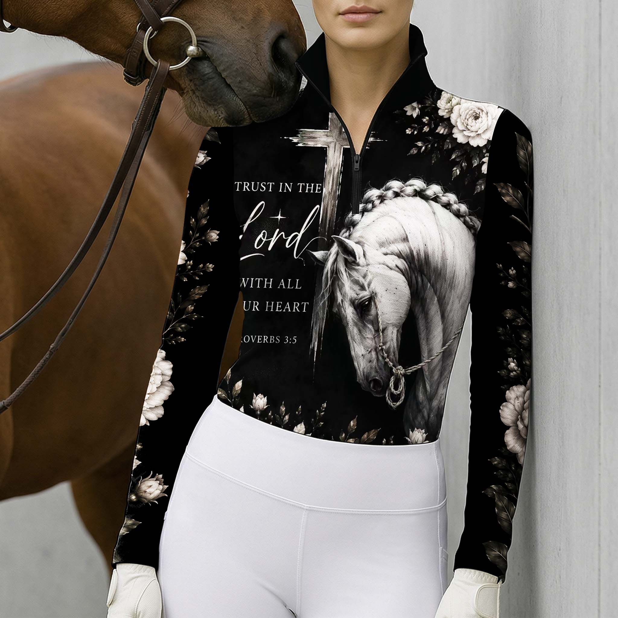 Model leaning against light grey stable wall beside chestnut horse wearing monochrome bible verse braided horse floral riding top
