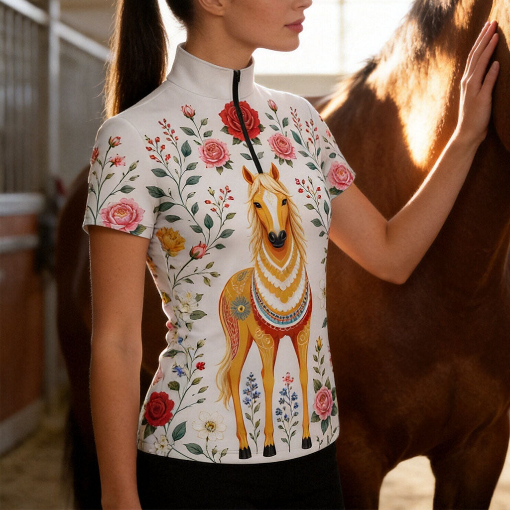 Barn stable lifestyle shot of female rider standing beside brown horse wearing white folk‑art golden‑horse and rose‑floral print short‑sleeve quarter‑zip riding top