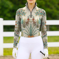 Model wearing the cream version of the Folk Art Horse & Botanical Print Long-Sleeve Riding Top at an outdoor equestrian arena, with jumps in the background