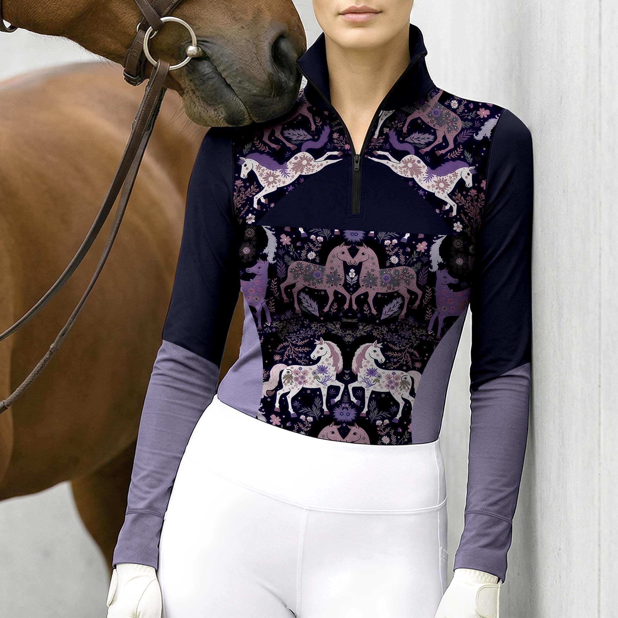 Model leaning against light grey stable wall beside chestnut horse wearing dark purple floral folk horse raglan long sleeve riding top
