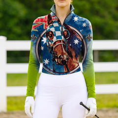 Woman wearing RIDER ATELIER Australian Racehorse Top at a sunlit pasture with a white fence, holding a riding crop, showing the full front print and green-to-blue gradient sleeves
