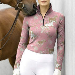 Outdoor lifestyle shot of female rider in dusty‑rose‑pink long‑sleeve quarter‑zip riding top with galloping‑horse wild‑floral print beside brown horse