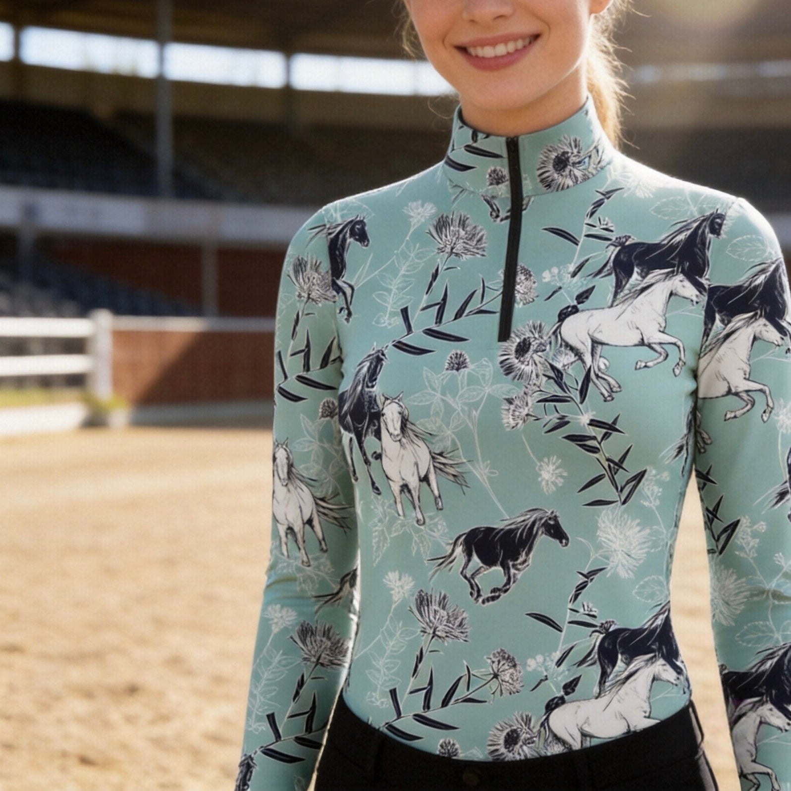Equestrian arena model shot of rider wearing muted sage teal long‑sleeve riding top with all‑over horse‑and‑wildflower sketch print