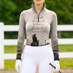 Full-body model shot wearing minimalist grey faith equestrian long sleeve top at sunlit green pasture white wooden fence, holding riding crop
