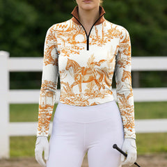 Outdoor fence‑side lifestyle shot of female equestrian wearing white‑base long‑sleeve riding top with orange Halloween toile‑de‑jouy graveyard‑horse all‑over print