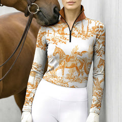 Stable‑wall lifestyle shot of female equestrian beside horse wearing white‑base long‑sleeve riding top with orange Halloween toile‑de‑jouy graveyard‑horse all‑over print