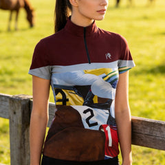 Model wearing the Horse Racing Finish Print Riding Top at a sunlit pasture, leaning on a wooden fence
