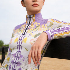 Model wearing lavender floral riding top leaning on dark wooden fence at sunlit pasture