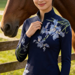 Lifestyle shot of female model leaning against wall with horse, wearing lavender watercolor iris floral long‑sleeve quarter‑zip riding top