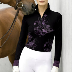 Lifestyle shot of female rider wearing black lavender lily butterfly horse line‑art long‑sleeve riding top beside brown horse