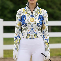 Full-body model shot wearing bright coastal tile faith equestrian long sleeve top at sunlit green pasture white wooden fence, holding riding crop
