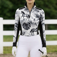 Full-body model shot wearing slimming black white tropical equestrian riding top at sunlit green pasture white wooden fence, holding riding crop
