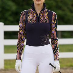 Full-body model shot wearing refined earth-tone abstract purple bloom gold swirl color block long sleeve top at outdoor equestrian show jumping arena with spectator stands, holding riding crop
