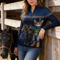Wooden stable fence lifestyle shot of female rider in navy cowboy‑cat‑and‑black‑pony print quarter‑zip riding top