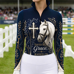 Model standing beside brown horse’s head wearing deep navy Saved By Grace cross white horse gold foliage horse bit long sleeve riding top against light grey stable wall
