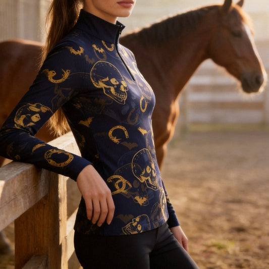 Outdoor golden‑hour pasture fence lifestyle shot of female equestrian wearing navy long‑sleeve quarter‑zip riding top with golden skull bat horseshoe all‑over print