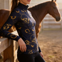 Outdoor golden‑hour pasture fence lifestyle shot of female equestrian wearing navy long‑sleeve quarter‑zip riding top with golden skull bat horseshoe all‑over print