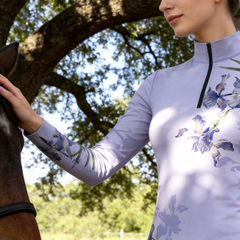 Golden sunlit riding‑arena lifestyle shot of model wearing navy iris floral long‑sleeve quarter‑zip riding top