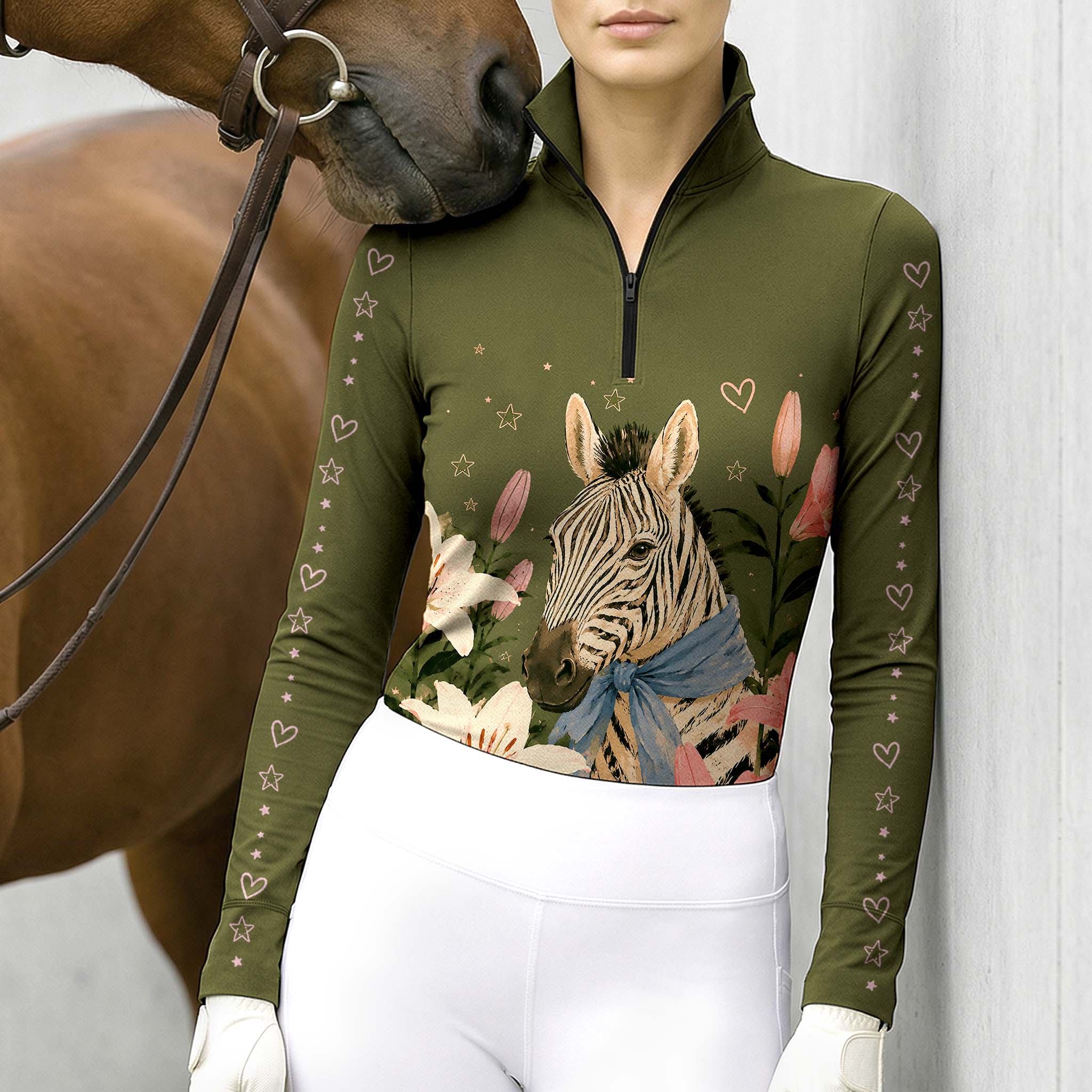 Stable‑side lifestyle shot of female equestrian beside horse wearing olive‑green long‑sleeve riding top with zebra lily floral and pink heart‑star sleeve print