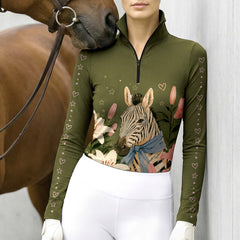 Stable‑side lifestyle shot of female equestrian beside horse wearing olive‑green long‑sleeve riding top with zebra lily floral and pink heart‑star sleeve print