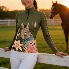 Golden‑hour outdoor pasture lifestyle shot of female equestrian leaning on white fence wearing olive‑green long‑sleeve quarter‑zip riding top with zebra lily floral and pink heart‑star sleeve print