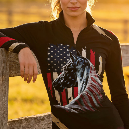 Woman wearing RIDER ATELIER black American flag horse quarter-zip top at sunset leaning on a wooden fence