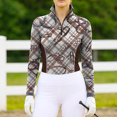 Woman wearing the RIDER ATELIER Brown Plaid & Equestrian Tack Print Long-Sleeve Quarter-Zip Riding Top at a sunlit pasture with a white fence, holding a riding crop, showing the print’s warm tones in natural daylight

