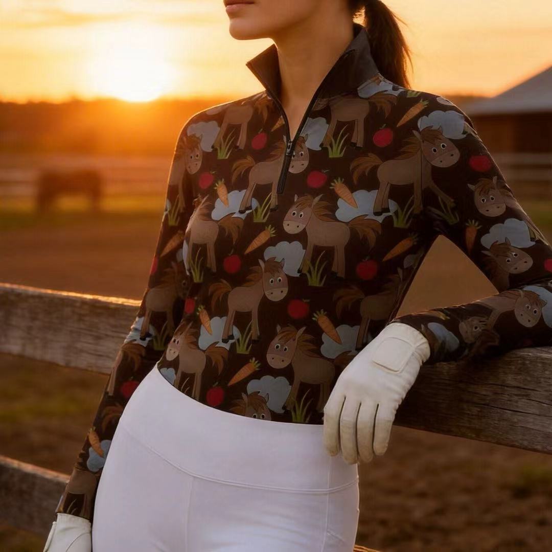Woman wearing RIDER ATELIER cute cartoon horse dark brown quarter-zip top leaning on a fence at sunset