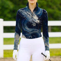 Woman wearing the RIDER ATELIER Deep Blue Chinese Dragon Print Long-Sleeve Quarter-Zip Riding Top at a sunlit pasture with a white fence, holding a riding crop, showing the print’s deep blue tones in natural daylight
