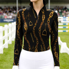 Model wearing the RIDER ATELIER Black Gold Chain & Stirrup Print Riding Top at an outdoor equestrian arena, with a white fence in the background
